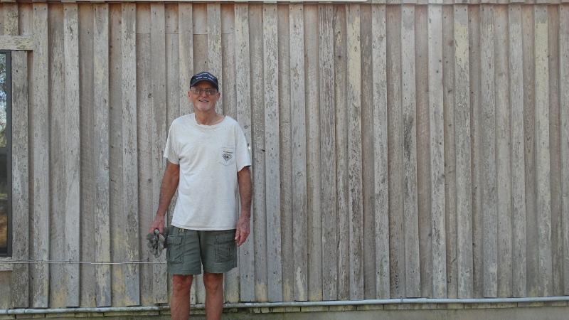 a man in work clothes stands against a wooden fence, below a sign reading "Troy Springs State Park"